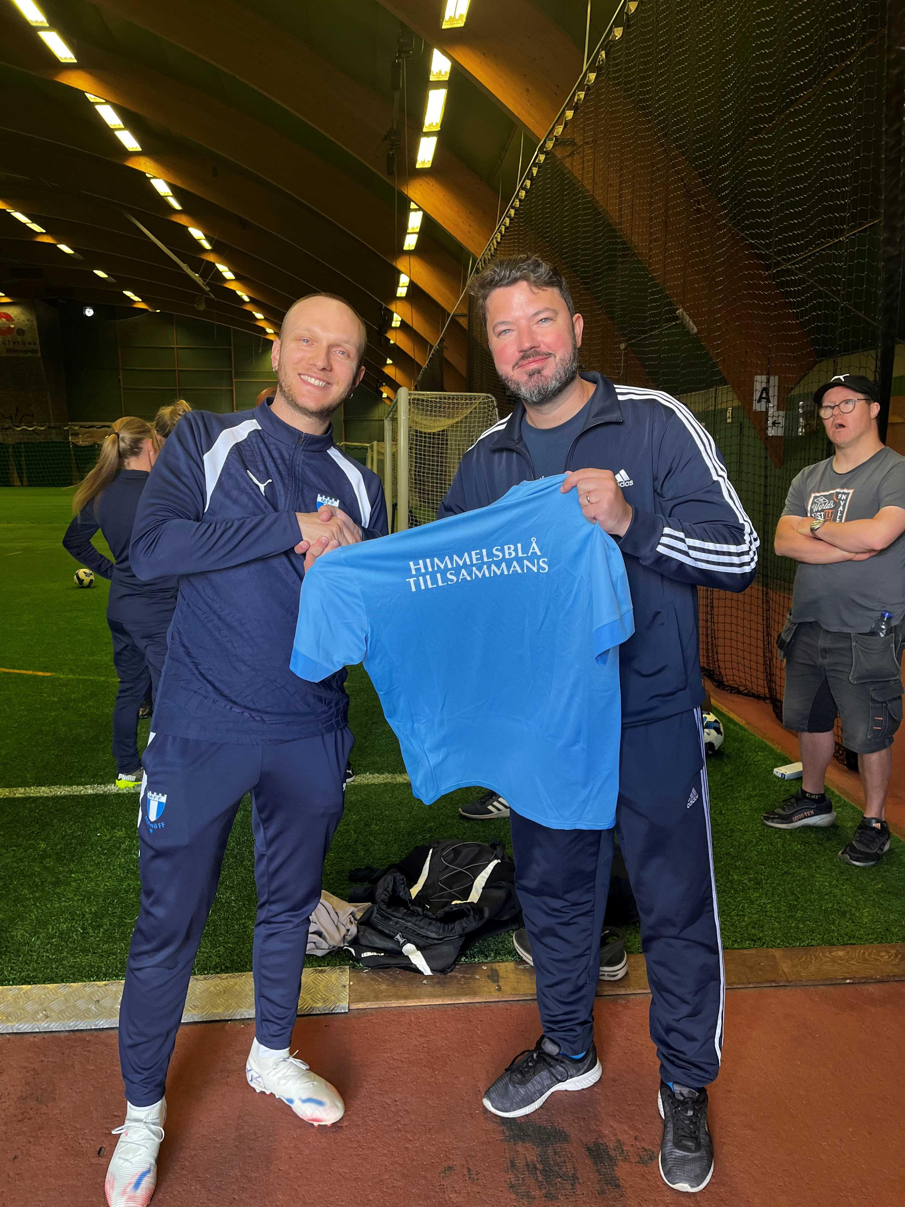 Luigi och Roko  håller i varsin sida av en blå tröja som det står Himmelsblå tillsammans på.