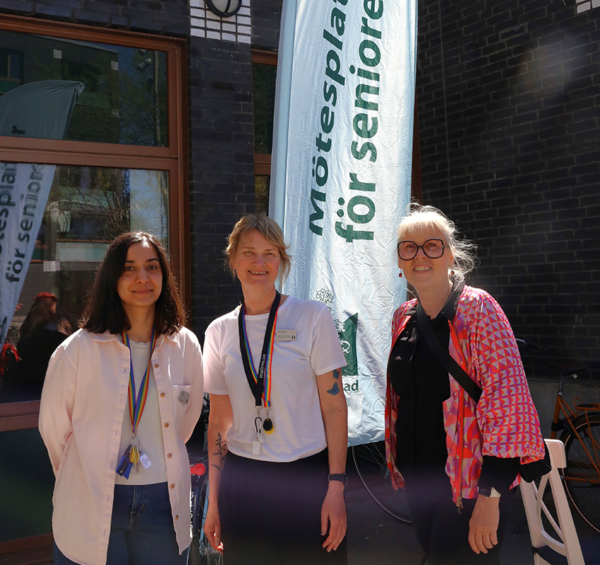 Patricia, Jenny och Elisabeth står tillsammans utanför mötesplatsen framför en en flagga där det står "Mötesplatser för seniorer".