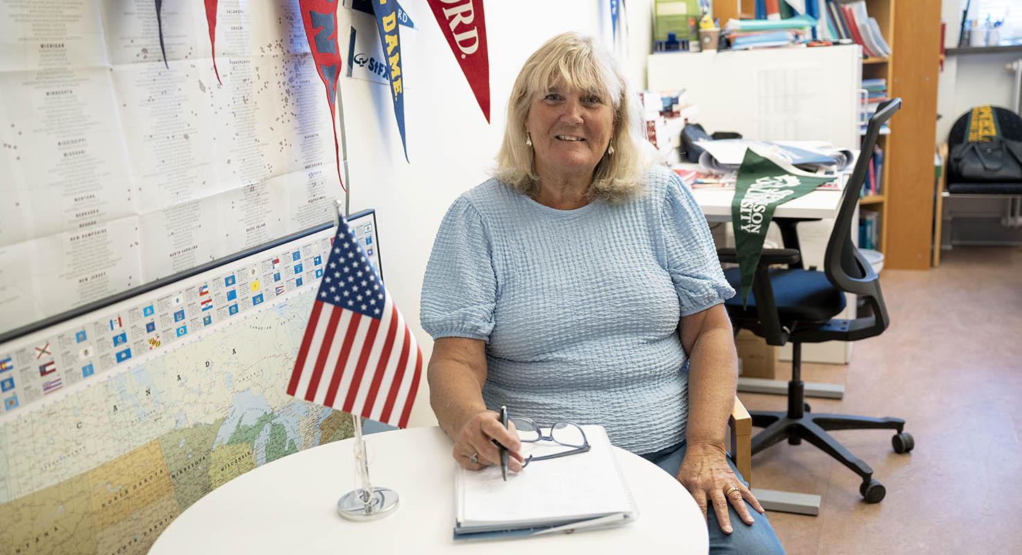 En kvinna sitter vid ett bord där amerikanska flaggan står. Bakom henne finns vimplar från många internationella universitet.