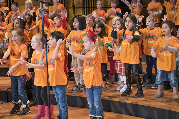 Massor med barn i orangea tröjor som sjunger för full hals. De står på en stor scen. 