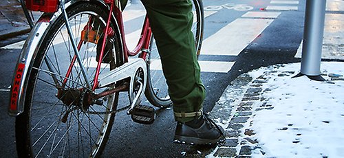 En cyklist står och väntar i en korsning. På marken ligger lite snö. 