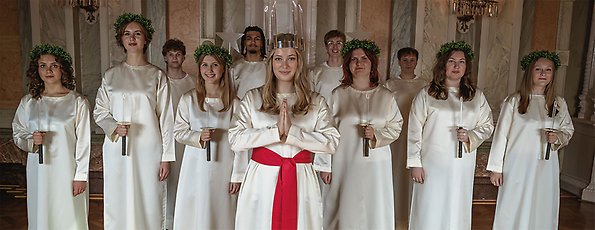 Malmö lucia 2025 med följe. Lucia och sex tärnor och fyra gossar står med ljus i händerna och ler mot kameran.