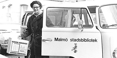Bibliotekarie stiger ur minibuss med väska som det står "Boken kommer" på.