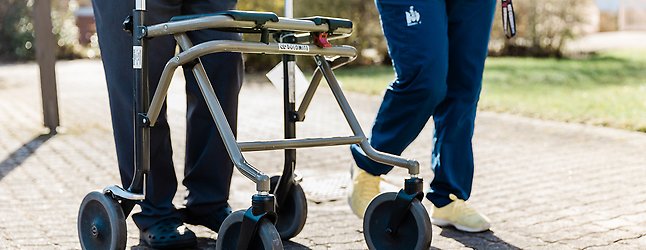 Äldre person med rullator på promenad med hemtjänstpersonal.
