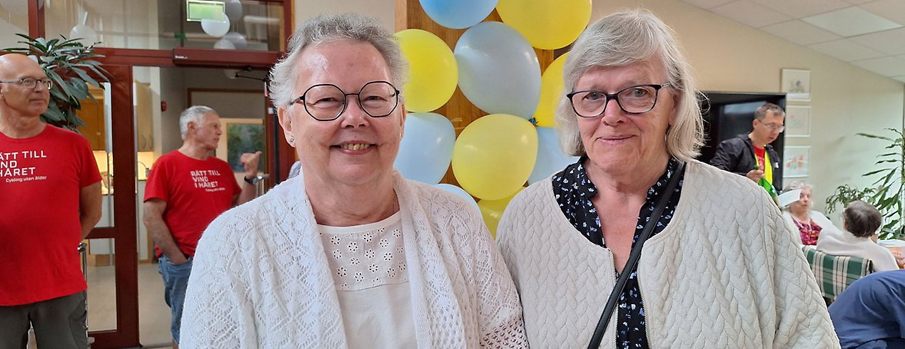 Två äldre kvinnor står framför ballonger. De har båda vita tröjor och glasögon.