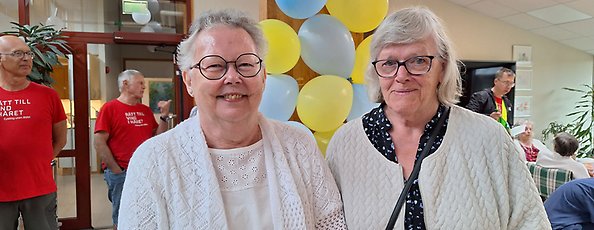 Två äldre kvinnor står framför ballonger. De har båda vita tröjor och glasögon.