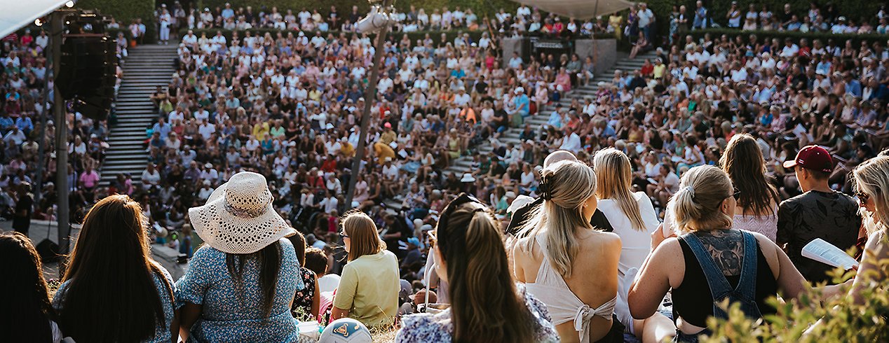 Stor samling människor sitter i solen och tittar på en konsert.