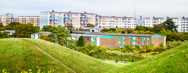 Holma Kroksbäck. Böljande gröna kullar i förgrunden, bostadshus i bakgrunden. 