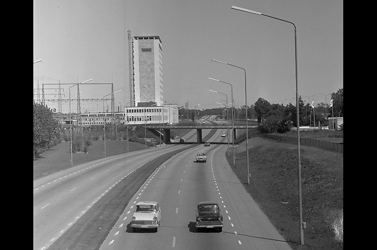 Svartvitt foto av motorväg, intill vägen längre fram i bild ligger ett ljust höghus.