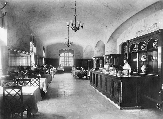 Svartvitt foto av restaurang med bord och stolar till vänster och en bardisk till höger.