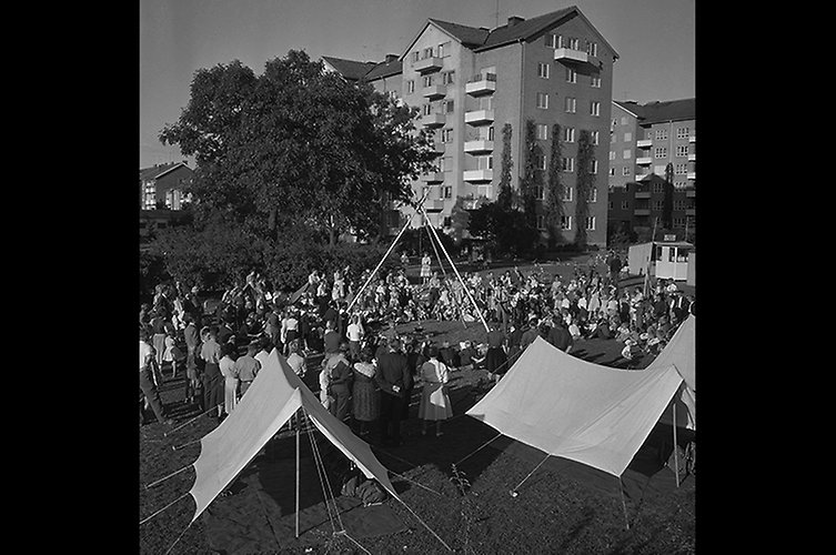 Svartvitt foto med uppspända vindskydd i förgrunden och massor av barn som sitter på gräset. I bakgrunden höga bostadshus.