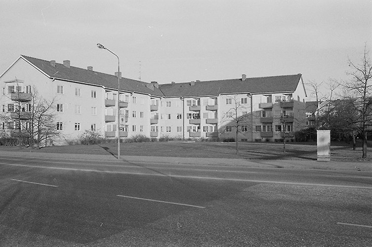 Svartvitt foto med en bred väg i förgrunden. I bakgrunden ett ljust flervåningshus i vinkel.
