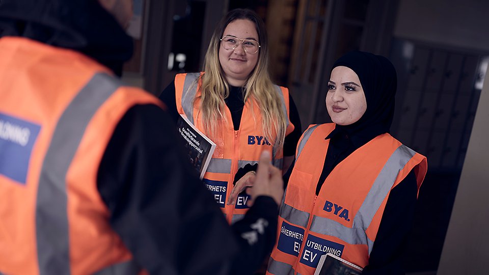 Två elever som läser till väktare står och samtalar med en tredje elev. Eleverna bär orange reflexväst med texten säkerhetsutbildning elev.