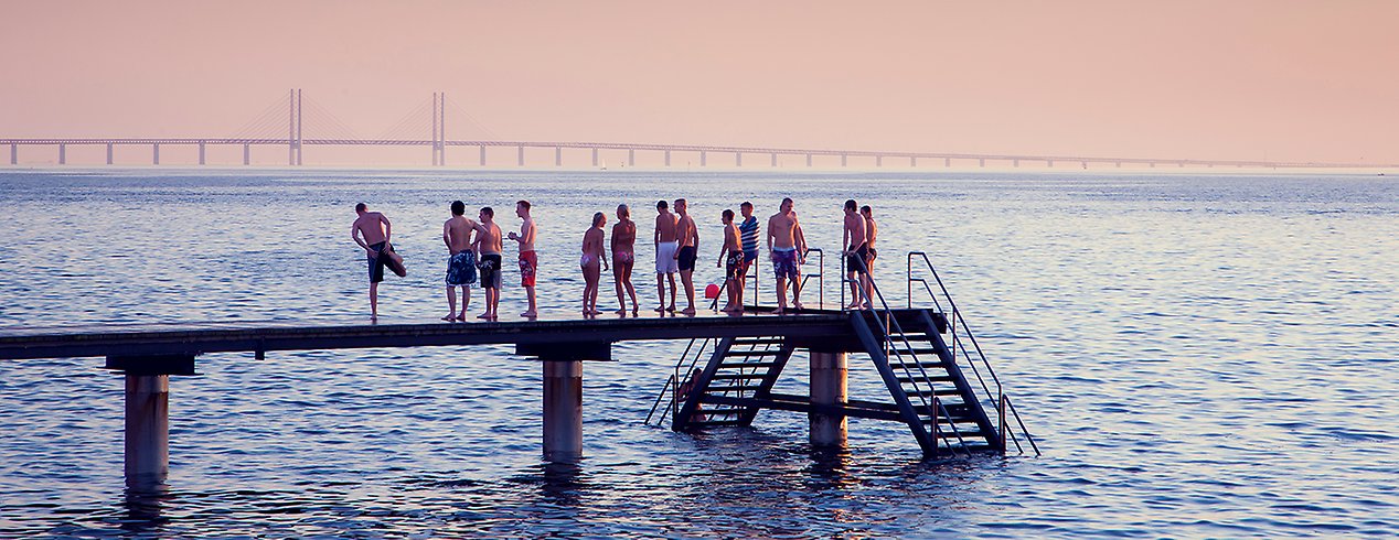 En samling ungdomar i badkläder står på en badbrygga, med Öresundsbron i bakgrunden.