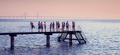 En samling ungdomar i badkläder står på en badbrygga, med Öresundsbron i bakgrunden.