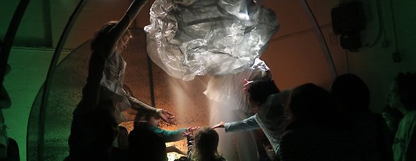 Flera personer barn och vuxna stå i ett rum ett molnformatobjekt hänger i luften, de sträcker sin hand mot det. 