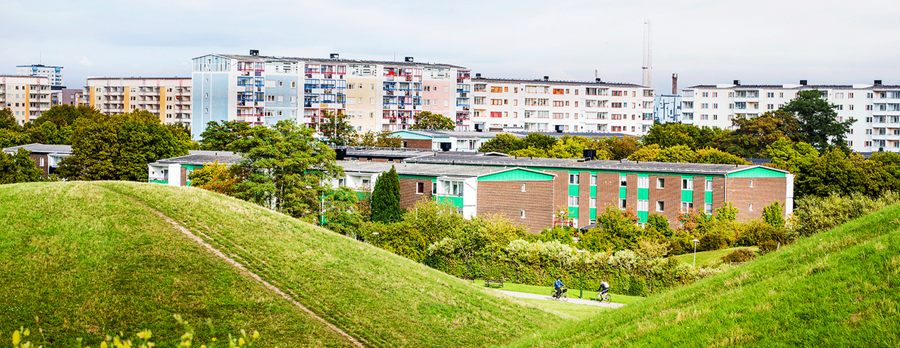 Holma Kroksbäck. Böljande gröna kullar i förgrunden, bostadshus i bakgrunden. 