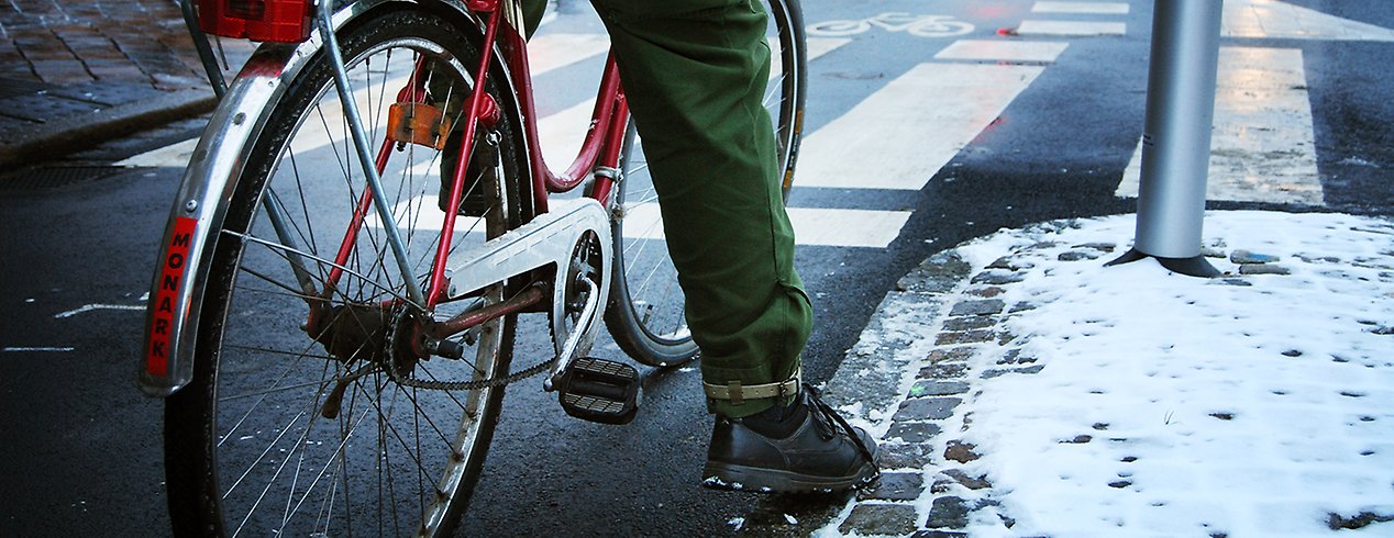 En cyklist står och väntar i en korsning. På marken ligger lite snö. 
