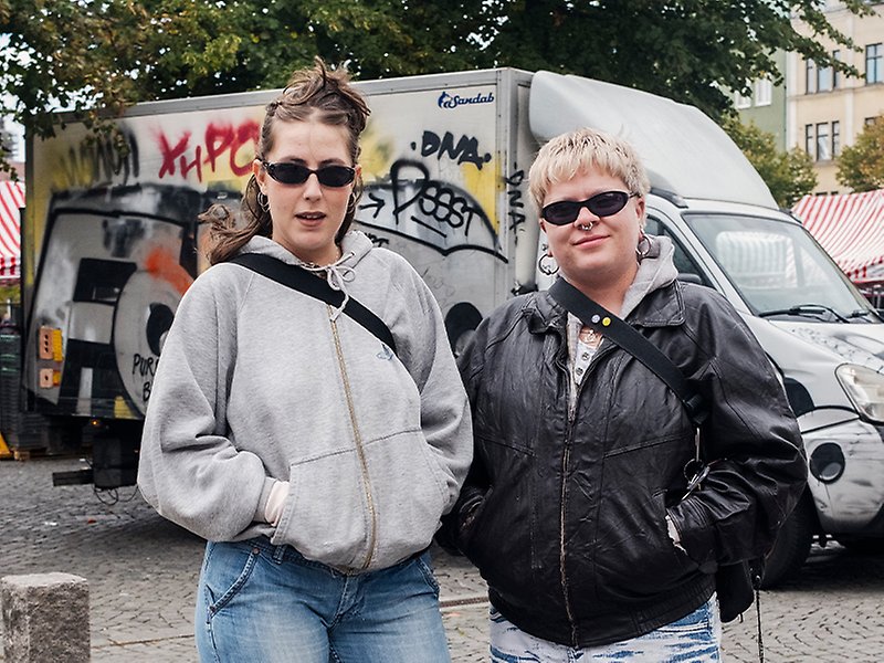 Två personer står på Möllevångstorget och poserar framför kameran. Den ena bär grå hoodtröja och den andra svart skinnjacka.