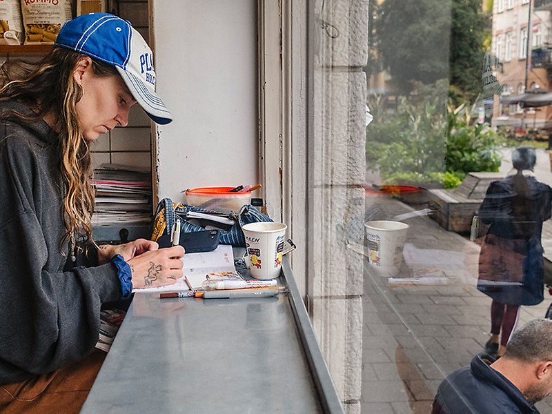 Konstnären Magda Lundberg sitter och arbetar på Kaffebaren. Genom fönstret syns en person sitta på uteserveringen och en annan gå förbi.