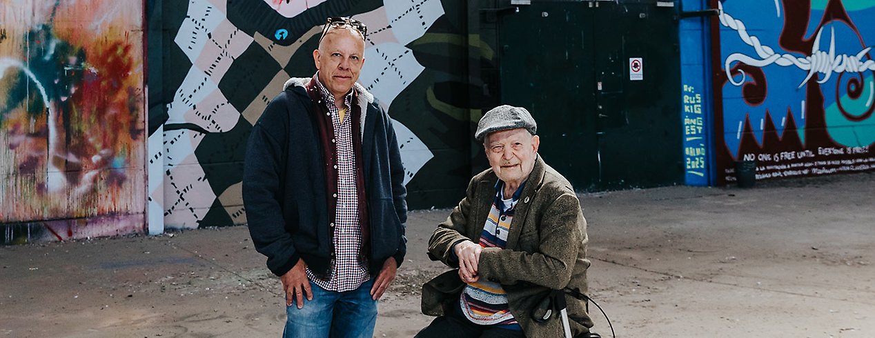 Pärra har en rutig skjorta och blå jacka och står bredvid Göte som sitter på sin rollator. Han har en grå keps och brun kavaj.