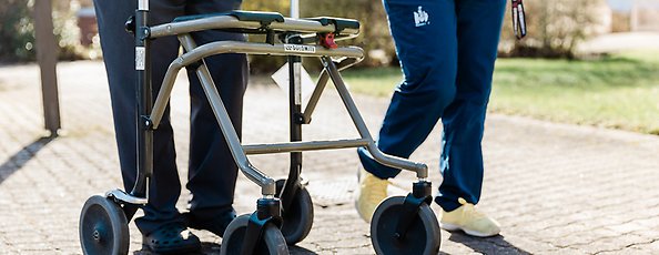 Äldre person med rullator på promenad med hemtjänstpersonal.