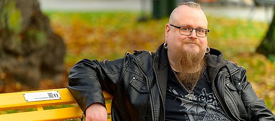 Man med skägg, glasögon, stora ringar i öronen och svarta kläder ler in i kameran.