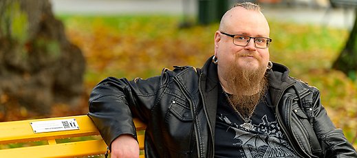 Man med skägg, glasögon, stora ringar i öronen och svarta kläder ler in i kameran.