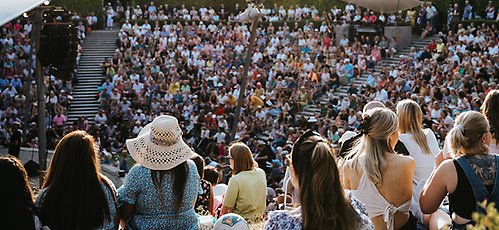 En stor samling människor sitter och tittar på en konsert. Solen skiner.