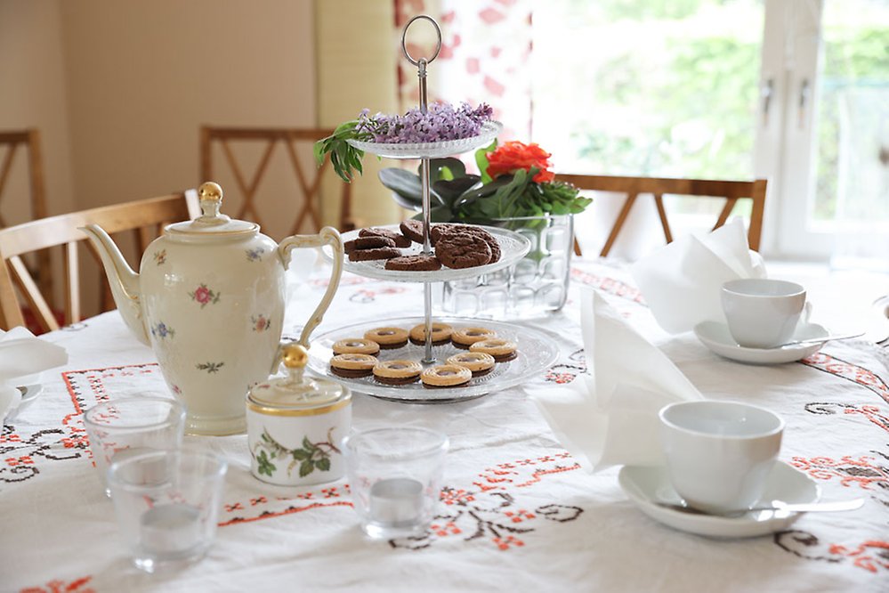 Ett uppdukat bord med linneduk, kakor och finporslin bjuder in till fika.