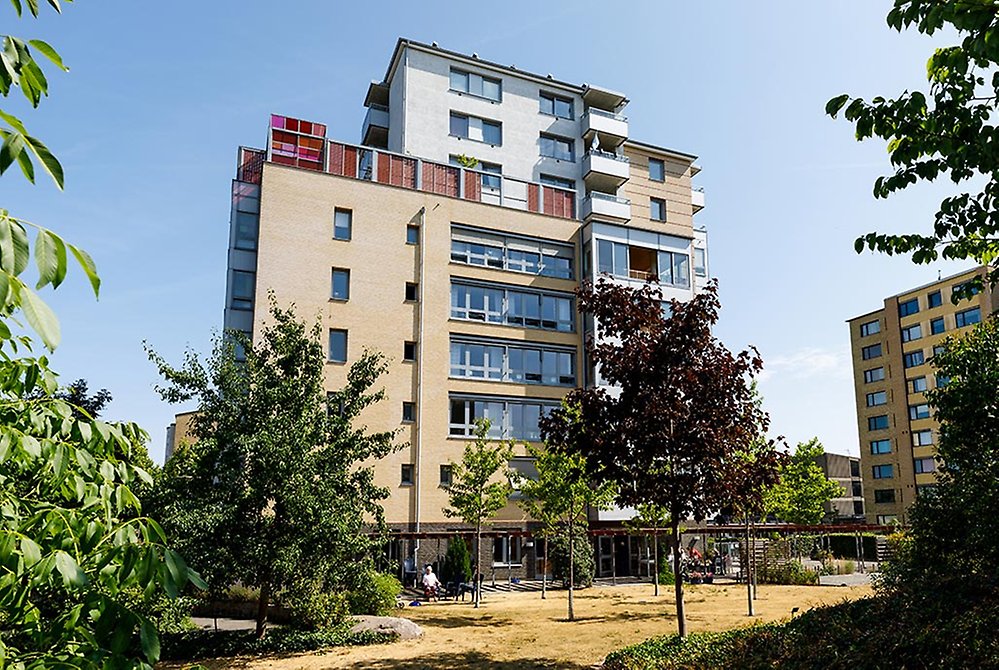 Huset är högt och man ser en färgglad terrass. På marken
framför huset finns en park med flertalet träd.