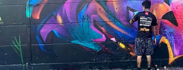 En person i svart t-shirt står vid en vägg och målar en muralmålning på väggen, målningen har olika färgnyanser i blått, rosa, orange och rött.