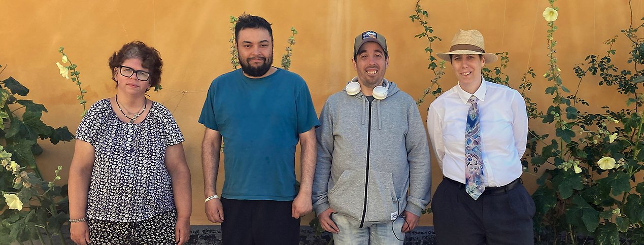 Fyra personer står framför en gul fasad och stockrosor med ljusgula blommor och ler mot kameran