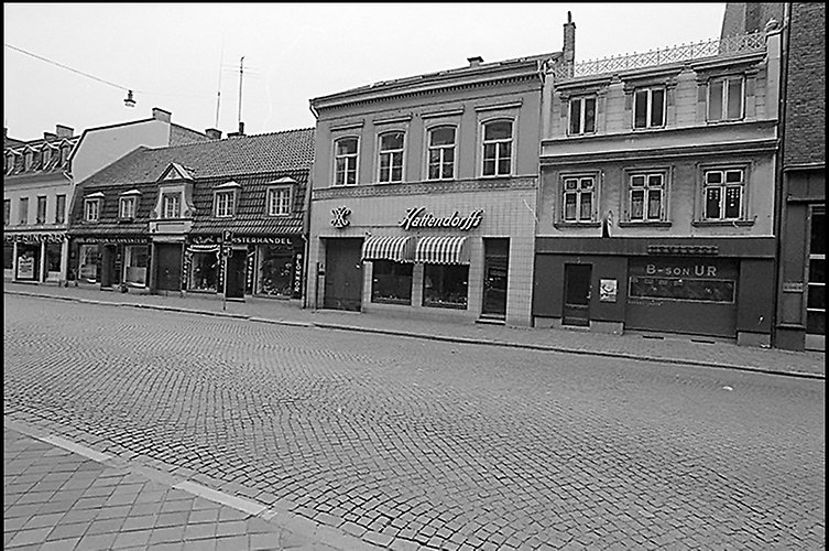 Kullerstensgata med fyra tvåvåningshus med olika fasader, butikslokaler i bottenvåningen.
