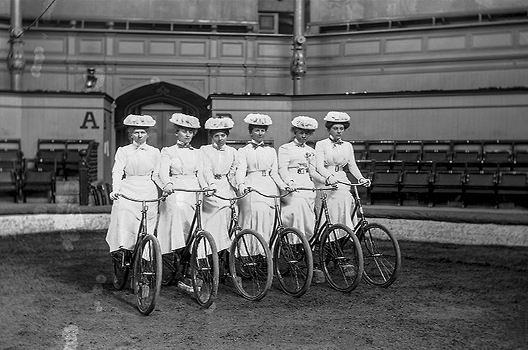 Sex kvinnor i dräkt poserar med sina cyklar i cirkuslokalen. 