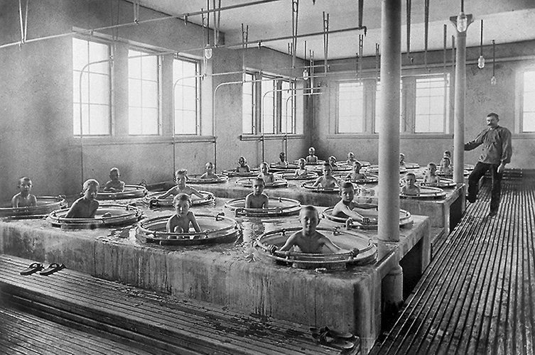 Pojkarna badar en och en i varsitt runt kar, karen står i raka rader och en manlig lärare övervakar badandet.