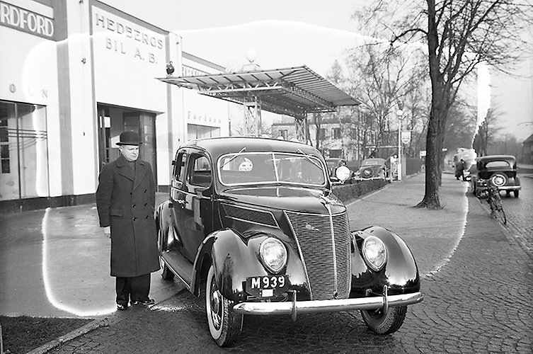 Äldre svartvitt fotografi av personbil med man bredvid