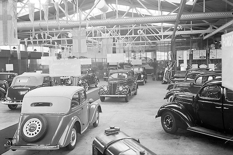 Äldre svartvitt fotografi. En mängd bilar till försäljning i en hangar. 