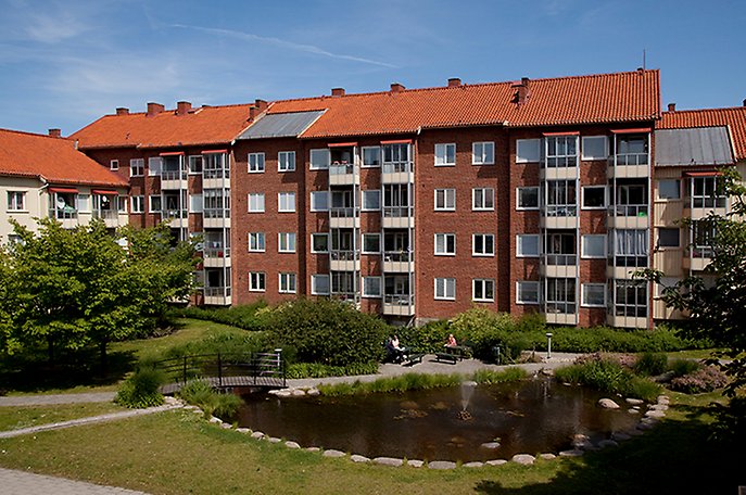 Bostadshus i rött tegel i bakgrunden, i förgrunden en damm och gröna ytor.