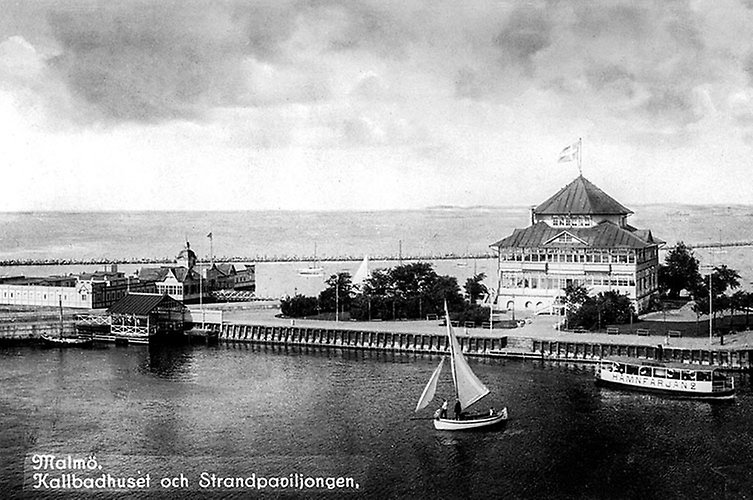 Färja och segelbåt framför hus med ett torn högst upp.