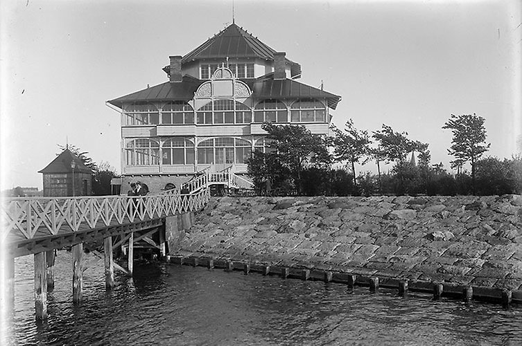 Brygga och kaj i förgrunden, hus med torn i bakgrunden.