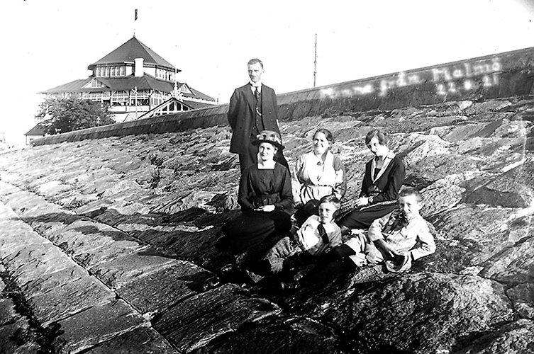 En man står upp på en kaj, tre kvinnor och två pojkar sitter ner framför honom. Ett hus med torn i bakgrunden.