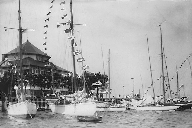 Flera vita segelbåtar i förgrunden. Flaggspel. Hus med torn i bakgrunden.