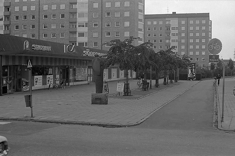 Rekangulärt torg med enplansbyggnad med butik och bank längs ena sidan. Två höga hus i bakgrunden.