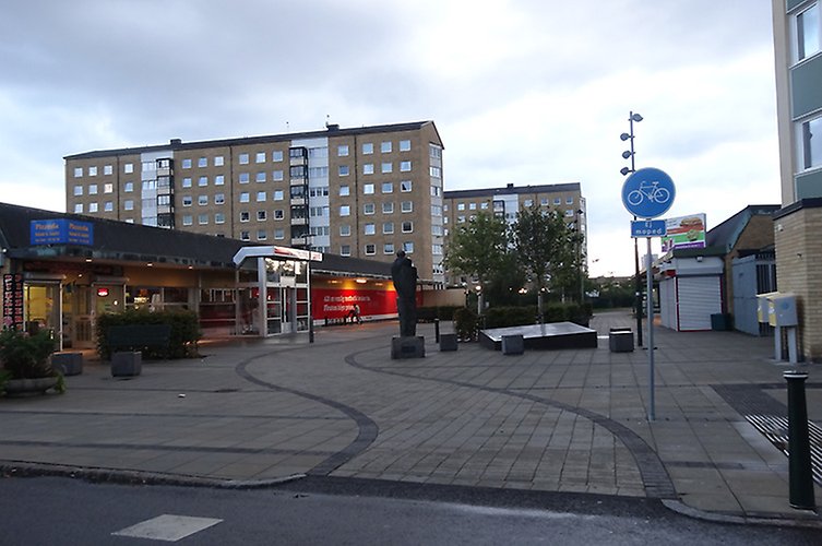 Rektandulärt torg med låg byggnad med butiker längs ena sidan. Två höga gula hus i bakgrunden.