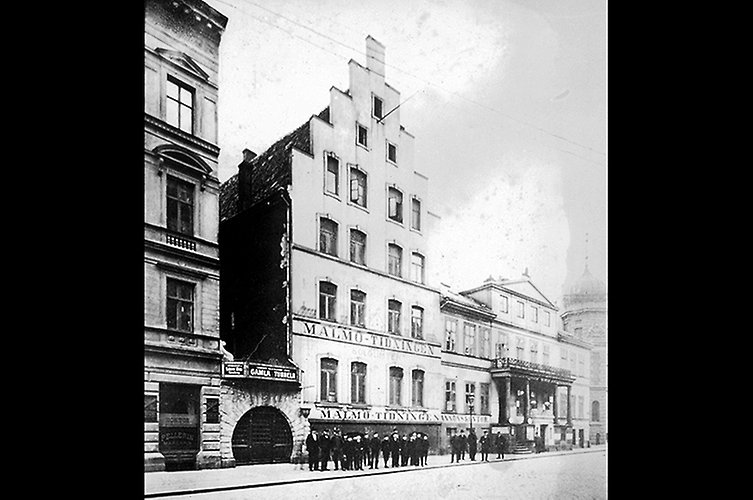 Tre äldre byggnader med gata i förgrunden. Svartklädda människor på trottoaren.
