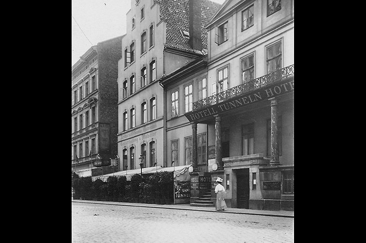 Tre hus i rad. Gata i förgrunden, en dam i hatt promenerar.