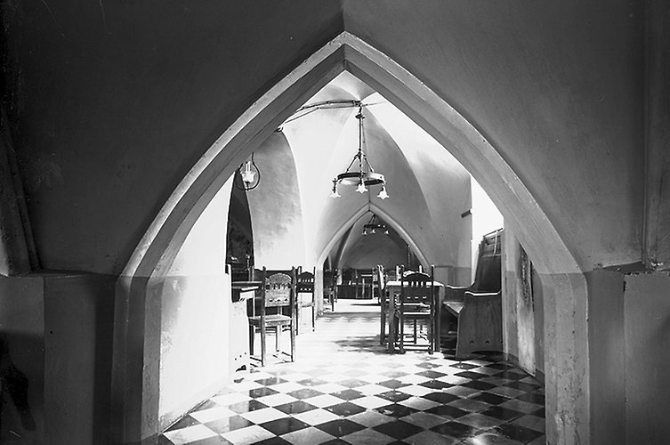 Spetsigt valv inomhus. Rutigt golv. Bord och stolar, en enkel takkrona i taket.