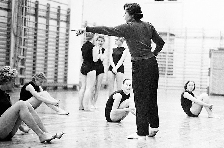 Kvinna instruerar flickor i gymnastikdräkt. Flickorna sitter på golvet i en gymnastiksal.