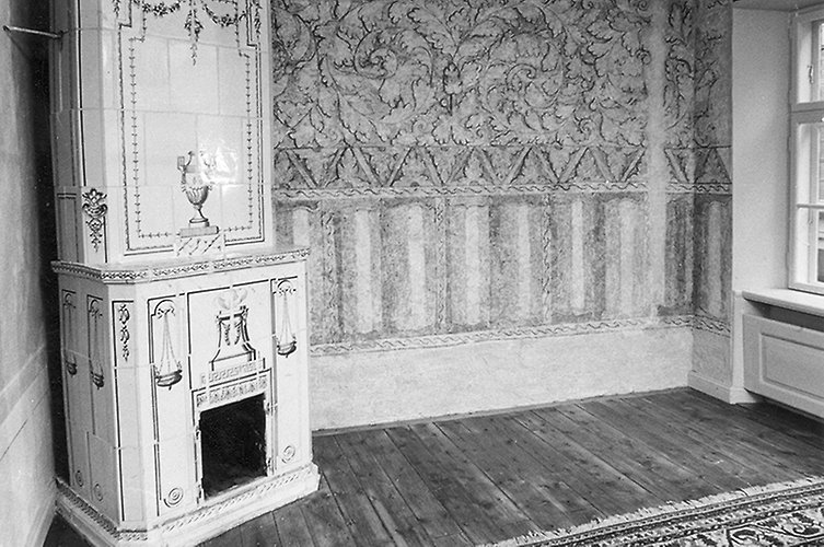 Äldre svartvitt foto på ett rum med kakelugn. Dekorationer på väggen.